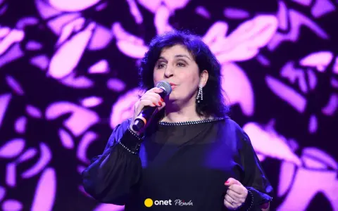 Kobieta śpiewa na scenie, w czarnej, ozdobnej sukni. Wiek męża Eleni nie jest widoczny na zdjęciu.