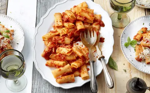 Ein Teller mit Rigatoni mit Tomatensauce, garniert mit Parmesan und Basilikum. Daneben ein Glas Weißwein.