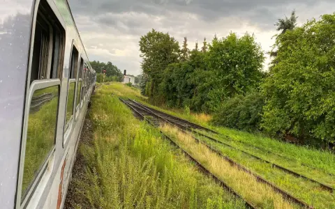 Widok z okna pociągu na tory kolejowe otoczone zielenią i chmury na niebie.