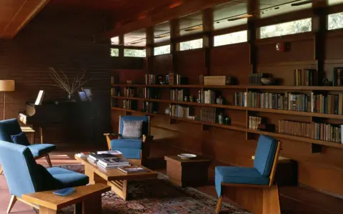 A cozy living room with blue chairs, a piano, and bookshelves. The clerestory windows above let in natural light, illuminating the warm wood interior.