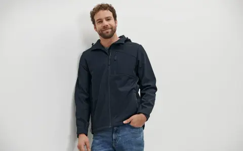 Mann mit lockigem Haar trägt eine dunkelblaue Softshelljacke und Jeans. Perfekt für das nächste Abenteuer oder zum Softshelljacke waschen.