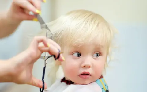 Ein kleines blondes Kind bekommt die Haare geschnitten. Die Schere nähert sich vorsichtig den feinen Strähnen.