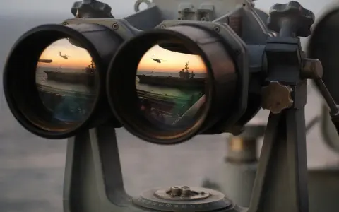 Fernglas-Werte: Ein Flugzeugträger und ein Hubschrauber spiegeln sich im Okular eines Fernglases bei Sonnenuntergang.
