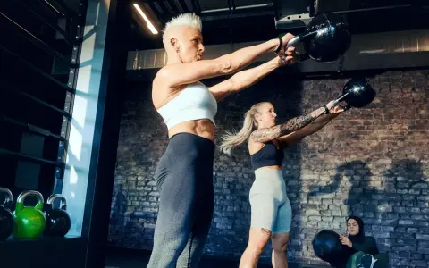 Ein Paar trainiert mit Kettlebells im Fitnessstudio. Sie heben Gewichte, während eine dritte Person im Hintergrund sitzt.