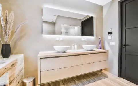 Elegantes baños modernos pequeños con lavabos dobles sobre mueble de madera clara, espejo iluminado y pared texturizada.