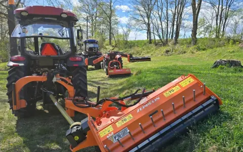Pomarańczowy mulczer używany, podłączony do traktora, gotowy do pracy na zielonej łące.