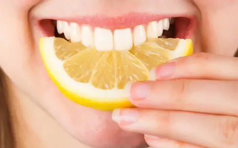 Une personne tient une tranche de citron près de sa bouche ouverte, révélant des dents blanches. Le citron est connu pour ses bienfaits sur la peau.