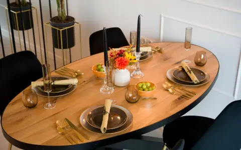 Un dîner élégant est prêt, avec des assiettes sombres, des couverts dorés et des raisins verts. Les art de la table sont mis en valeur par des bougies noires et des fleurs colorées.