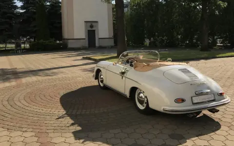 Biały, zabytkowy kabriolet Porsche, udekorowany kwiatami, czeka przed kościołem. To idealny samochod na wesele, gotowy na podróż poślubną.
