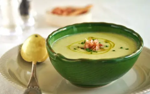 Un refrescante gazpacho de melón, adornado con jamón serrano y cebollino, servido en un cuenco verde con una cuchara dorada.