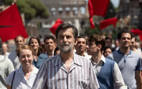 Nanni Moretti cammina in una folla con bandiere rosse, sullo sfondo il Colosseo.