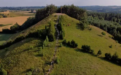 Wzgórze porośnięte trawą i drzewami, ze schodami prowadzącymi na szczyt, gdzie stoi drewniany krzyż. Widok na pola i lasy.