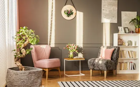 Un angolo relax con due poltrone, un tavolino con fiori e libri, una pianta e una libreria. La luce del sole crea un'atmosfera accogliente.