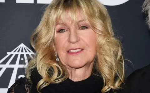Christine McVie, en ikonisk sångerska i Fleetwood Mac, ler med sitt blonda hår och smycken.