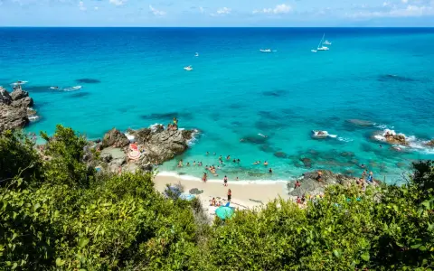 Spiaggia affollata con gente che nuota e prende il sole, barche in mare e vegetazione lussureggiante. Un angolo di paradiso del **calabria mare**.