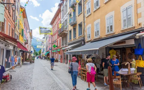 Rue commerçante animée à Aubenas-les-Alpes, avec des façades colorées, des drapeaux et des passants profitant de l'été.
