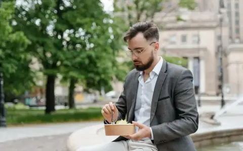 Mężczyzna w okularach i marynarce robi sobie przerwę w pracy, jedząc sałatkę na ławce w parku.