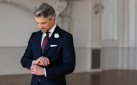 Elegancki mężczyzna w granatowym garniturze i białej koszuli, z bordowym krawatem i butonierką, przygotowuje się na wesele.