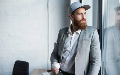 Mężczyzna w szarej czapce snapback i garniturze trzyma piłkę baseballową.