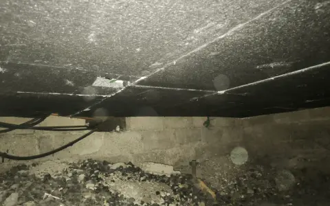 Vue de dessous d'un plancher, montrant le matériau d'isolation du plancher bas, des tuyaux et des blocs de béton.