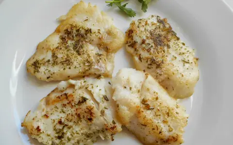 Cuatro trozos de pescado blanco rosada a la plancha, sazonados con hierbas, servidos en un plato blanco con una ramita de perejil.