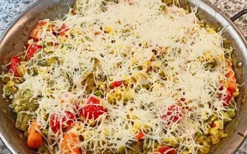 Arroz a la milanesa listo para hornear, cubierto con queso rallado y vegetales frescos como tomates, zanahorias y guisantes.