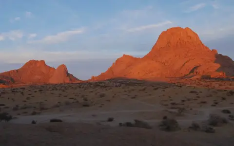 Zachód słońca maluje na pomarańczowo skalne szczyty Spitzkoppe w Namibii. Pustynny krajobraz z suchymi krzewami i ścieżkami.