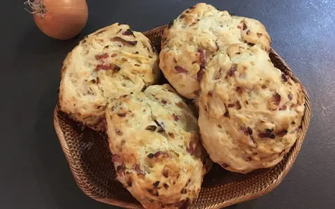 Quatre pains aux lardons et oignons dorés dans un panier en osier, avec un oignon cru à côté.