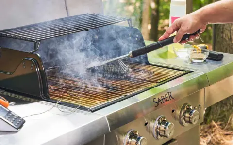 A hand uses a grill brush to clean the grates of a smoking Saber grill, showing the best way to clean a grill for your next cookout.