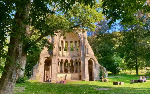 Ruinen einer alten Kirche in NRW, umgeben von Bäumen. Menschen genießen ihre Freizeit auf der grünen Wiese.