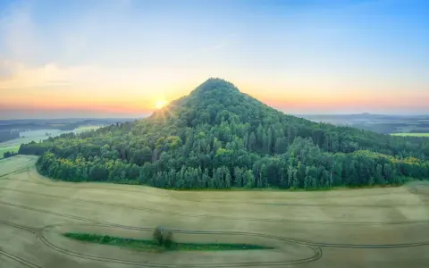 Góry Kaczawskie o wschodzie słońca. Stożkowate wzgórze porośnięte lasem, otoczone polami zbóż.