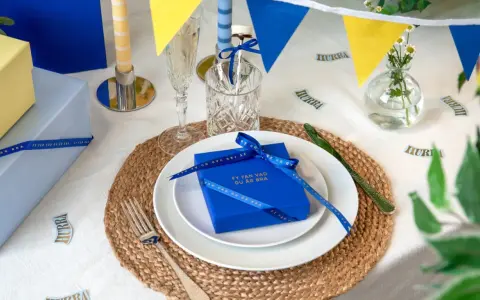 Blå och gula girlanger, ljus och presenter på ett dukat bord. En blå present med band och texten "Fy fan vad du är bra" är en fin pom pom dekoration.