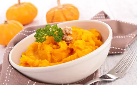 Un tazón de cremoso **puré de calabaza al horno**, adornado con perejil y nueces. Al fondo, calabazas pequeñas y una servilleta marrón.