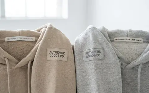 Dwa bluzy z kapturem, beżowa i szara, z naszywką "AUTHENTIC GOODS CO." i metką "100% ORGANIC COTTON | MADE IN JAPAN". Oryginalna metka Champion.