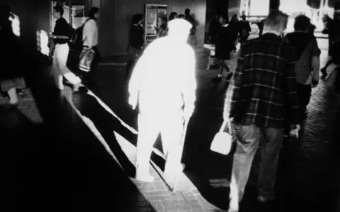 A stark black and white image by Trent Parke captures a blurred figure in a white hat and coat, walking with a cane. Other figures are also blurred, suggesting movement and a fleeting moment.