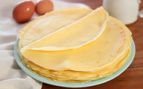 Piatto di crepes senza burro, leggere e dorate, con due uova sullo sfondo.