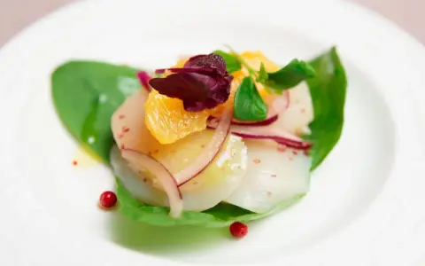 Carpaccio de saint jacques frais, garni d'orange, d'oignon rouge et de pousses vertes, sur une feuille d'épinard.