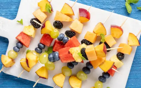 Spiedini di frutta colorati con pesche, melone, anguria, ananas, mirtilli, more e uva, guarniti con menta fresca.