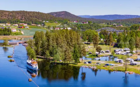 En båt på en sjö vid en campingplats i Norge. Husbilar och tält står utspridda på gräsmattor vid vattnet, omgivna av träd och kullar.