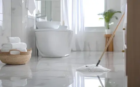 Czyszczenie łazienki w toku. Mop delikatnie przesuwa się po lśniącej podłodze, a stos białych ręczników czeka w wiklinowym koszu.