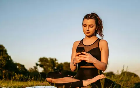 Kvinna i lotusställning på en matta utomhus, med en mobiltelefon i händerna. En modern typ av meditation.