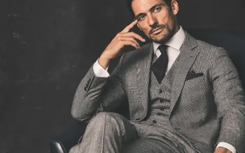 A man in a grey suit, black shirt, and tie, with a thoughtful expression, sits in a chair.