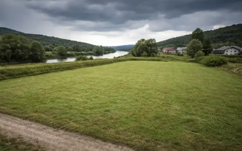 Łąka nad rzeką, domy w oddali. Chmury zapowiadają deszcz, budząc obawy o zagrożenie powodziowe.
