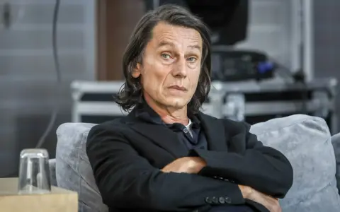 Krzysztof Warlikowski, reżyser teatralny, w czarnym garniturze, siedzi na kanapie. Jego życie prywatne budzi zainteresowanie.