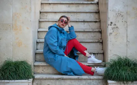 Kobieta w niebieskim płaszczu i czerwonych spodniach, w białych sneakersach, prezentuje sportową elegancję na kamiennych schodach.