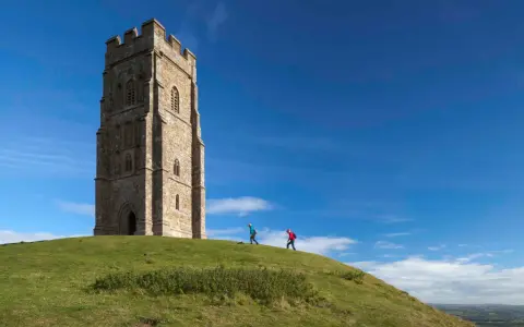 Two hikers ascend the grassy slope towards the iconic Glastonbury Tor tower under a bright blue sky.