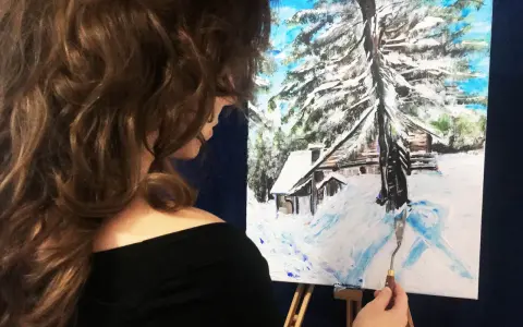 Artystka kończy malowanie zimowego domku w górach. Obraz przedstawia zasypany śniegiem krajobraz z chatką i drzewami.
