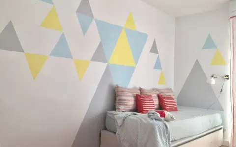 Pared pintada con triángulos de colores pasteles, creando un diseño geométrico moderno. La habitación tiene una cama con cojines y una manta.