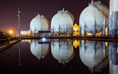 Duże, białe zbiorniki magazynujące gaz ziemny (natgas) odbijają się w wodzie. W tle nocne oświetlenie.