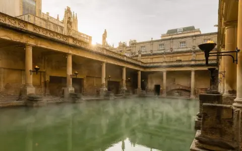Steaming green water fills the Great Bath, surrounded by ancient Roman ruins with columns and torches.